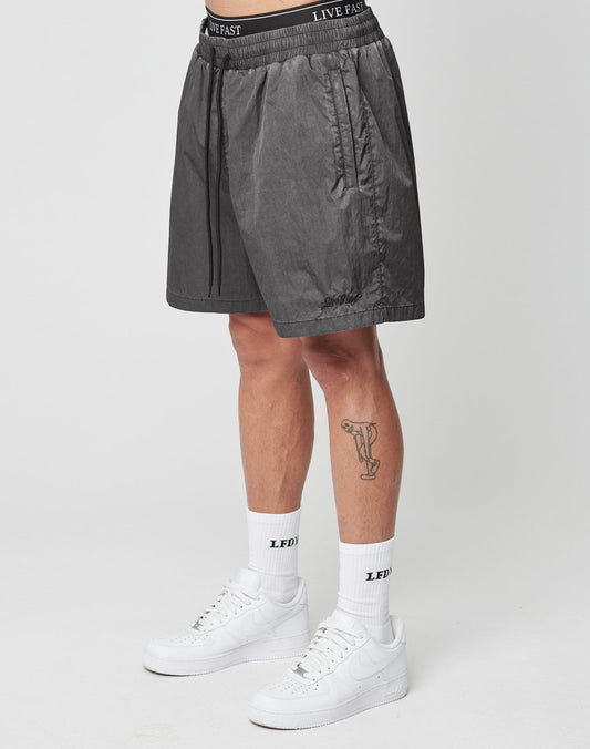 A person wearing LIVE FAST DIE YOUNGs black Live Fast Swimshorts, featuring a LIVE FAST waistband, stands against a white background. White LEDYF socks, white sneakers, and a leg tattoo finish the look. The shorts are made from polyamid.