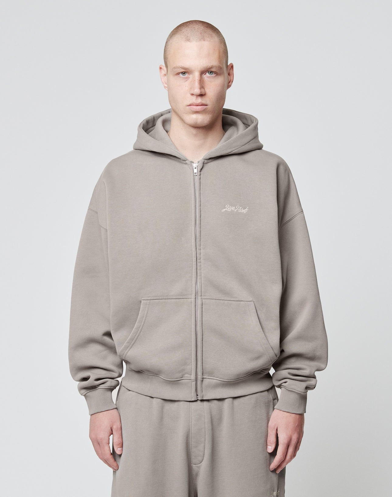 A person with a shaved head wears the LIVE FAST DIE YOUNG Basic Script Hooded Zip—a loose-fitting, gray Baumwolle-Polyester hoodie and sweatpants—standing against a light gray background, looking at the camera with a neutral expression.