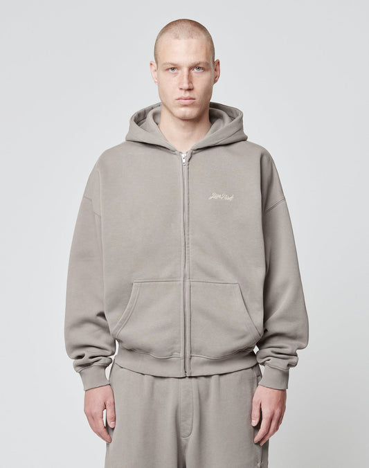 A person with a shaved head wears the LIVE FAST DIE YOUNG Basic Script Hooded Zip—a loose-fitting, gray Baumwolle-Polyester hoodie and sweatpants—standing against a light gray background, looking at the camera with a neutral expression.