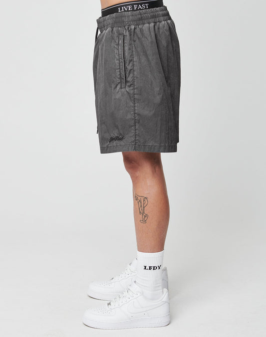A person stands sideways in tinted black, regular fit Live Fast Swimshorts by LIVE FAST DIE YOUNG with a branded waistband, white LFDY socks, and white sneakers. A leg tattoo is visible. Made from soft polyamide. Background: plain white.