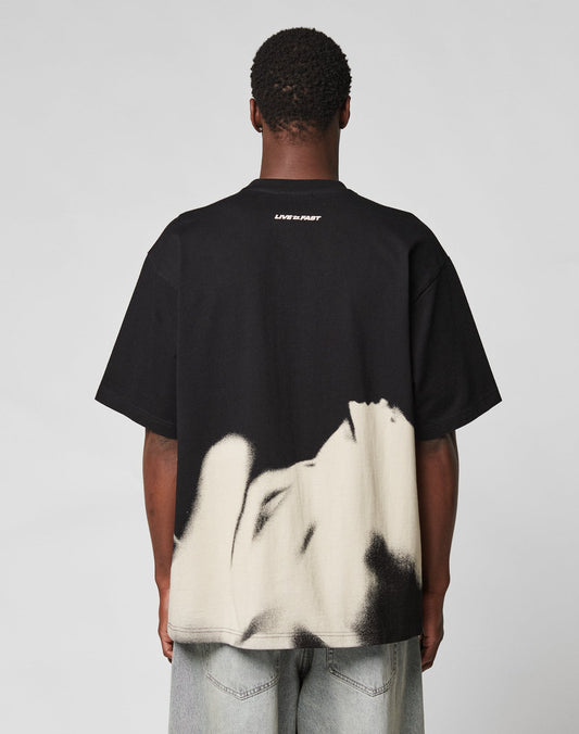 Someone faces away, wearing the LFDY Mystic Tee by LIVE FAST DIE YOUNG—an oversize black T-shirt made from 100% Baumwolle with bold front and back prints reading LIVE FAST—paired with light blue jeans.