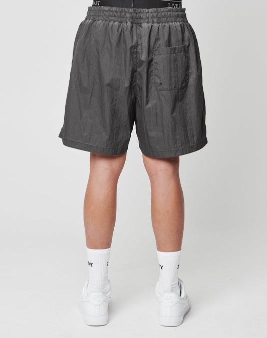 A person is seen from behind wearing LIVE FAST DIE YOUNG Live Fast Swimshorts in Tinted Black, featuring a regular fit, elastic waistband, and single back pocket, paired with white crew socks and sneakers on a plain white background.