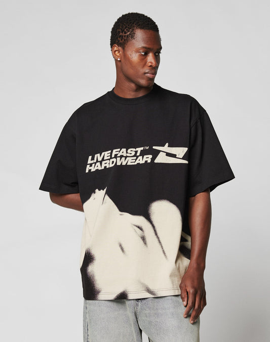 A man wears the LFDY Mystic Tee by LIVE FAST DIE YOUNG—an oversized black T-shirt with bold white LIVE FAST HARDWEAR front and back prints plus abstract graphics—paired with light gray jeans, against a plain background. Made of 100% Baumwolle.
