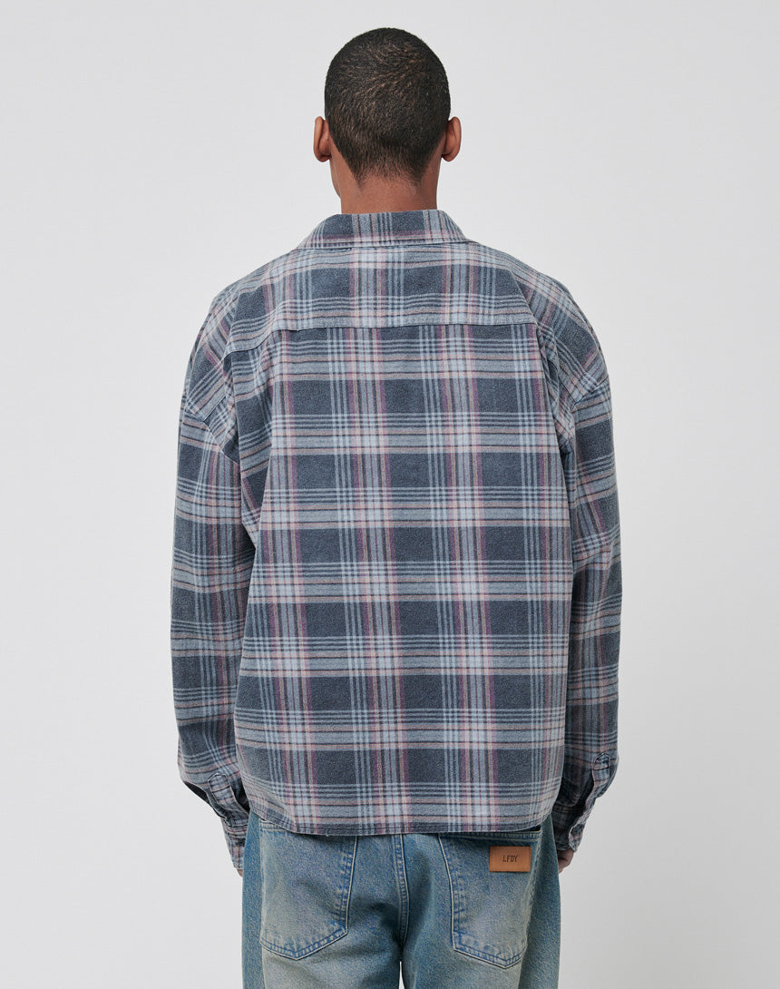 A person stands facing away, wearing the LIVE FAST DIE YOUNG Loose Flannel Shirt in blue, gray, and purple plaid (100% Baumwolle), paired with light blue jeans against a plain light background.