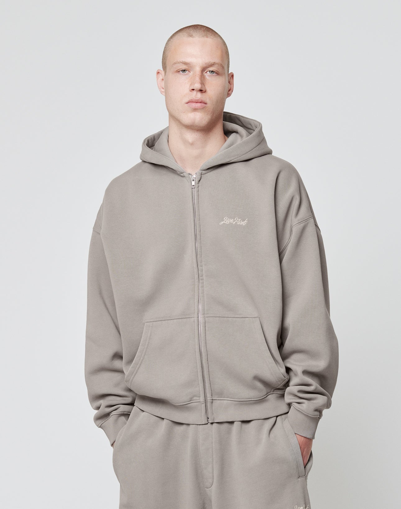 A person wearing the LIVE FAST DIE YOUNG Basic Script Hooded Zip in taupe with matching sweatpants stands against a plain light gray background, looking directly at the camera.