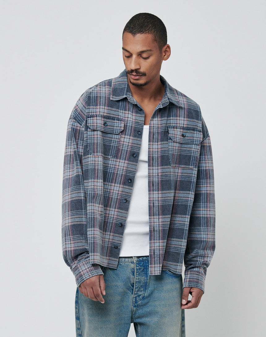A man in a LIVE FAST DIE YOUNG Loose Flannel Shirt with a vintage wash, layered over a white tee and blue jeans, stands against a plain light background, looking down with a neutral expression.