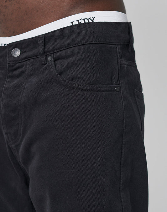 Close-up of a person wearing LIVE FAST DIE YOUNG Regular Denim jeans in black with a tapered leg, classic five-pocket style, and regular fit. A white waistband with partial LEDY text is visible above the jeans.