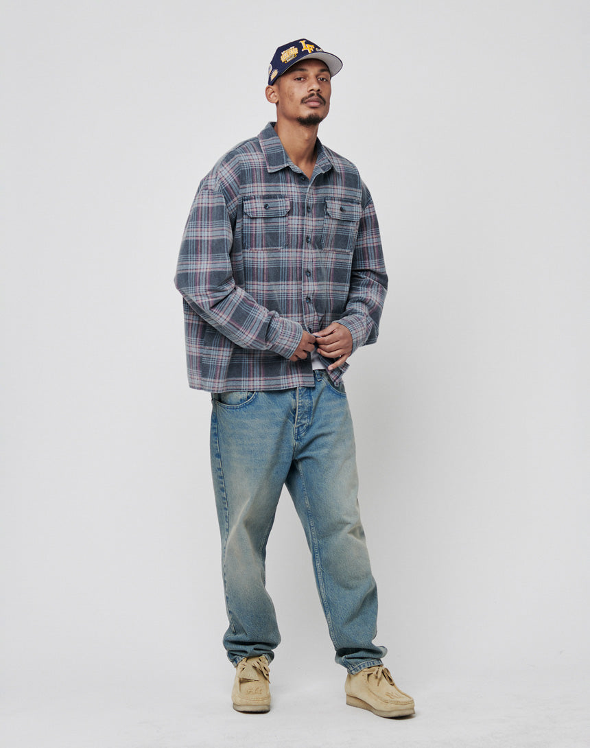 A man stands against a white background wearing the LIVE FAST DIE YOUNG Loose Flannel Shirt in gray and purple plaid, light blue vintage-wash jeans, tan shoes, and a dark baseball cap.