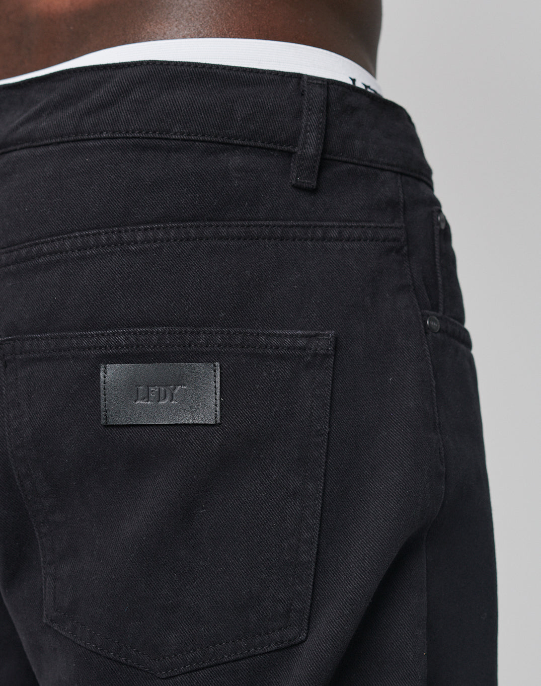 Close-up of a person wearing Regular Denim by LIVE FAST DIE YOUNG. Features a black LDY patch, tapered leg, regular fit, and back pocket detail. White underwear waistband is visible. Made from 100% Baumwolle for comfort.