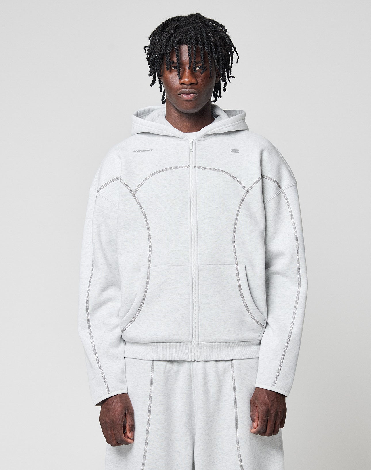 A person with short dreadlocks wears the LFDY Active Fleece Hooded Zip in Oversize Fit by LIVE FAST DIE YOUNG with matching sweatpants, standing against a plain light background and looking directly at the camera. | model