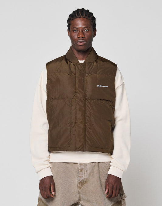 A man wears the LIVE FAST DIE YOUNG Live Fast Cropped Vest in brown over a cream sweatshirt and beige pants, standing against a plain light gray background.