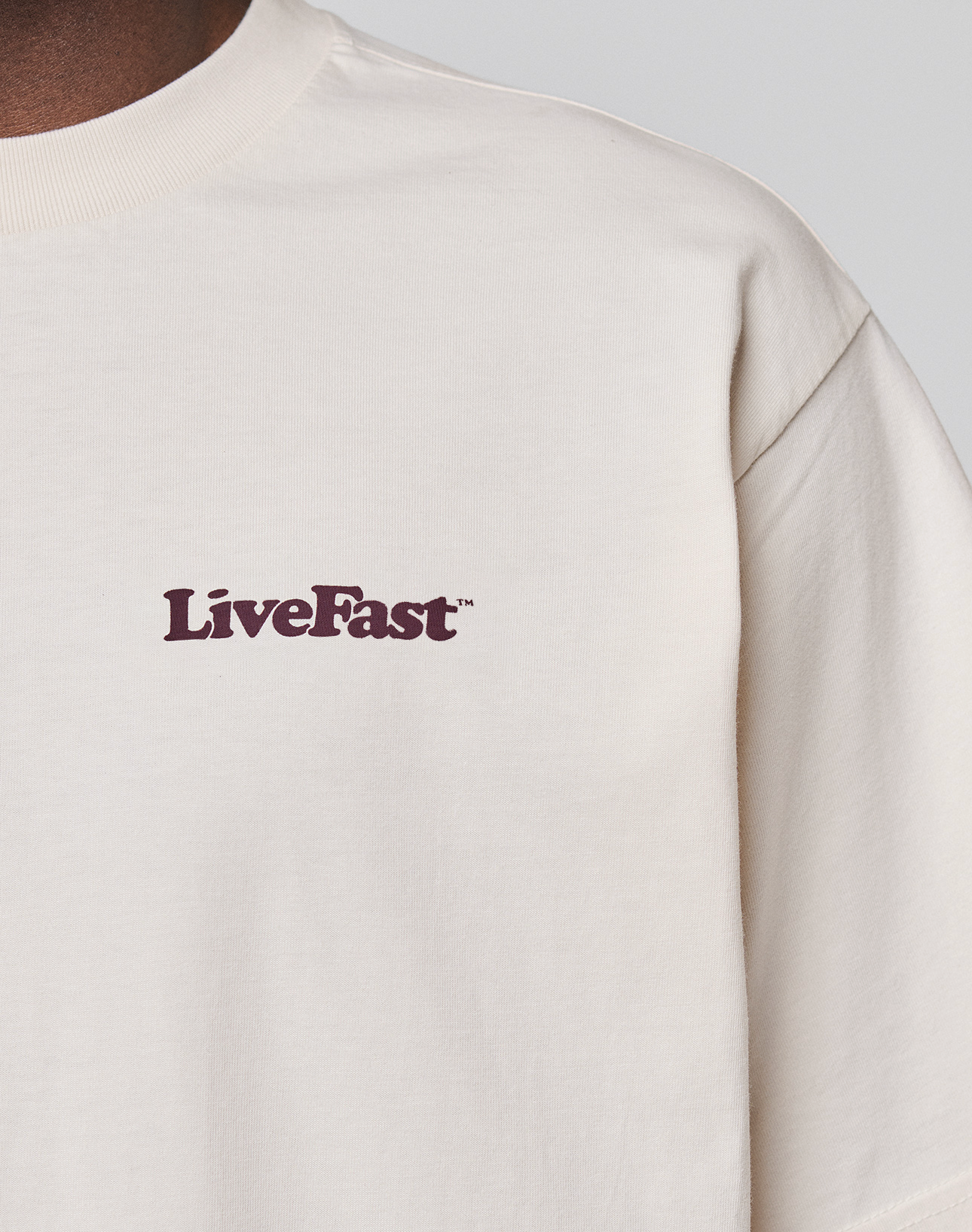 A close-up of a cream-colored Basic 210 Tee by LIVE FAST DIE YOUNG, featuring "LiveFast" in dark purple on the left chest. Only the upper torso, neck, and shoulder are visible. Made from 100% Baumwolle.