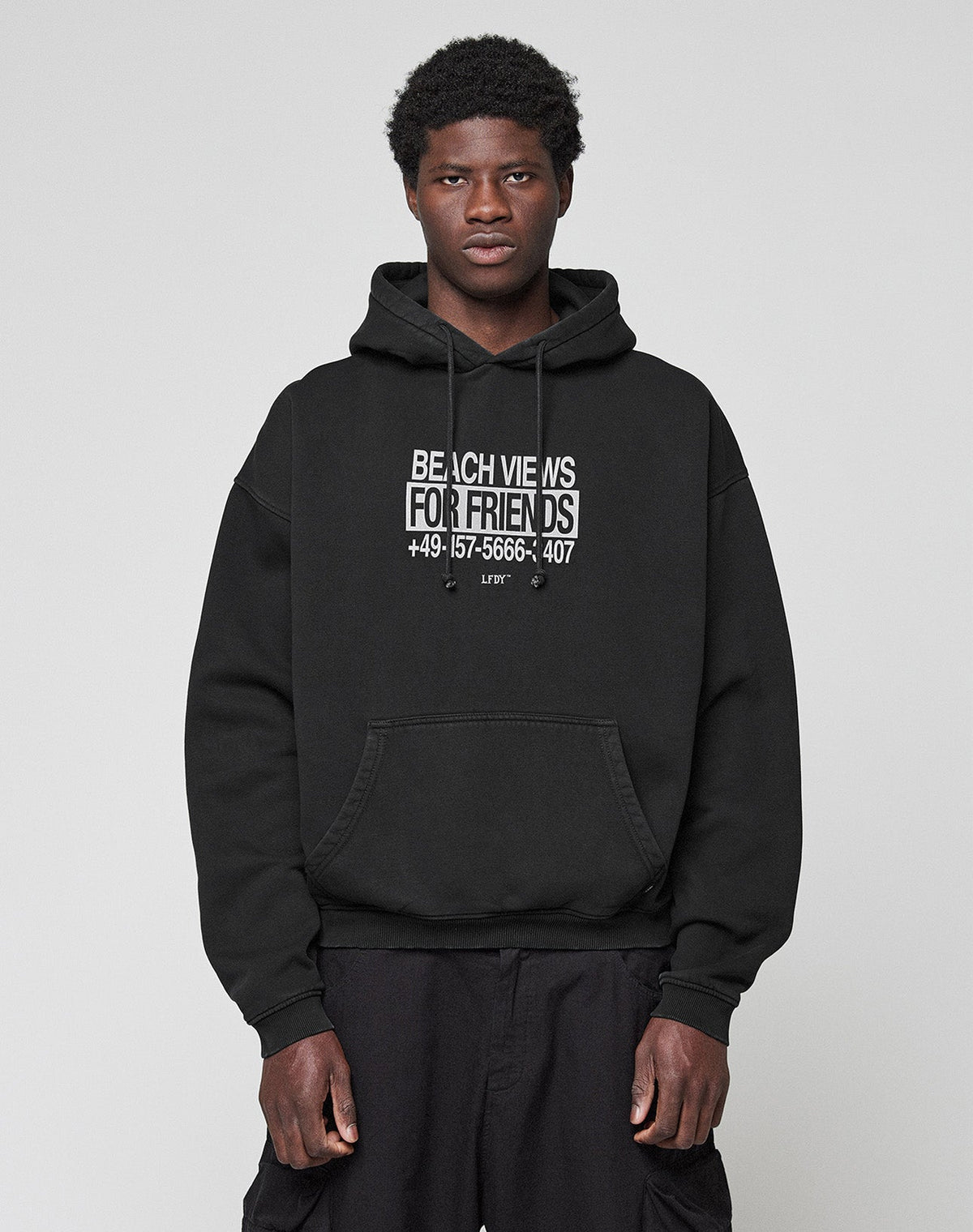 A person wears a black Live Fast Call Us Hooded by LIVE FAST DIE YOUNG, made of Baumwolle with BEACH VIEWS FOR FRIENDS +49-157-5666-3407 as bold front and back prints, standing against a plain light gray background.