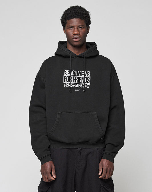A person wears a black Live Fast Call Us Hooded by LIVE FAST DIE YOUNG, made of Baumwolle with BEACH VIEWS FOR FRIENDS +49-157-5666-3407 as bold front and back prints, standing against a plain light gray background.