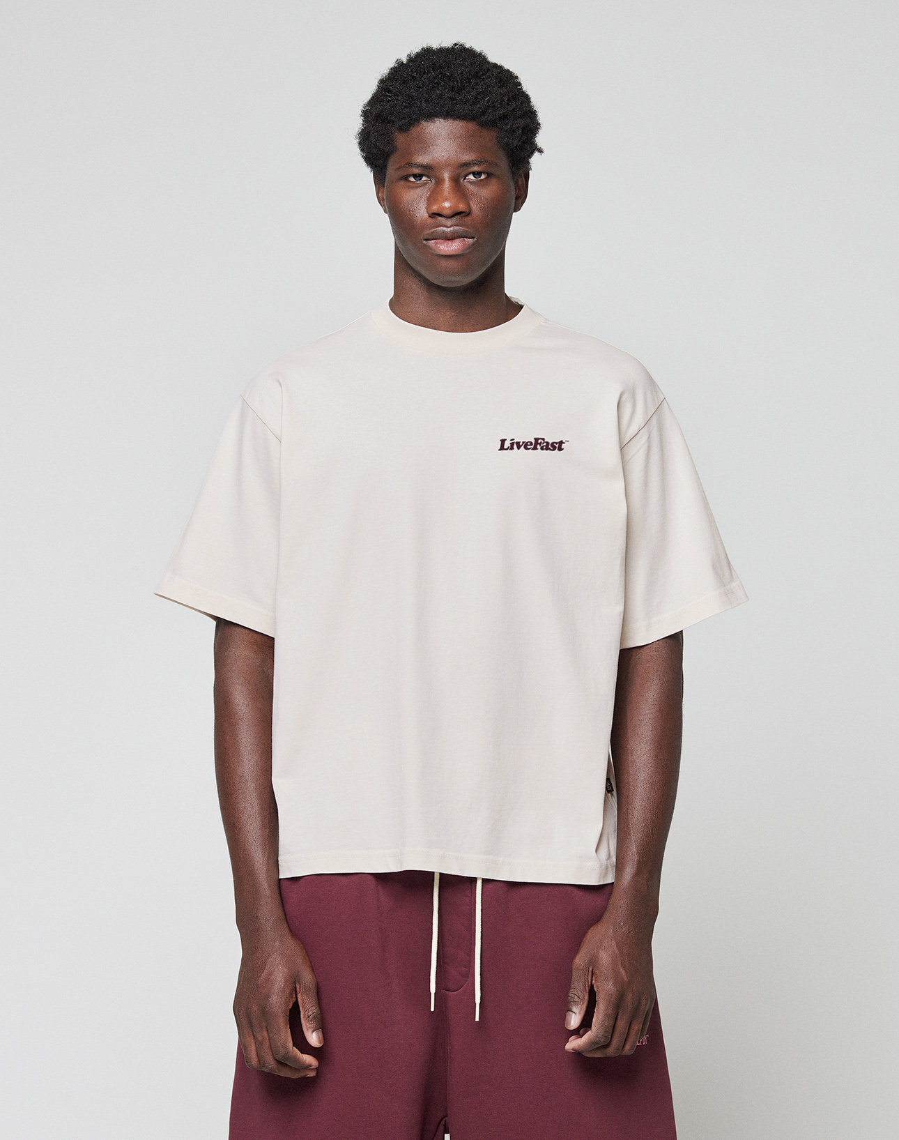 A person stands against a plain background wearing the LIVE FAST DIE YOUNG Basic 210 Tee in loose, light beige oversize fit with a LiveFast front print and maroon shorts. Made from 100% Baumwolle. Their arms rest at their sides as they look at the camera.