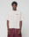 A person stands against a plain background wearing the LIVE FAST DIE YOUNG Basic 210 Tee in loose, light beige oversize fit with a LiveFast front print and maroon shorts. Made from 100% Baumwolle. Their arms rest at their sides as they look at the camera.