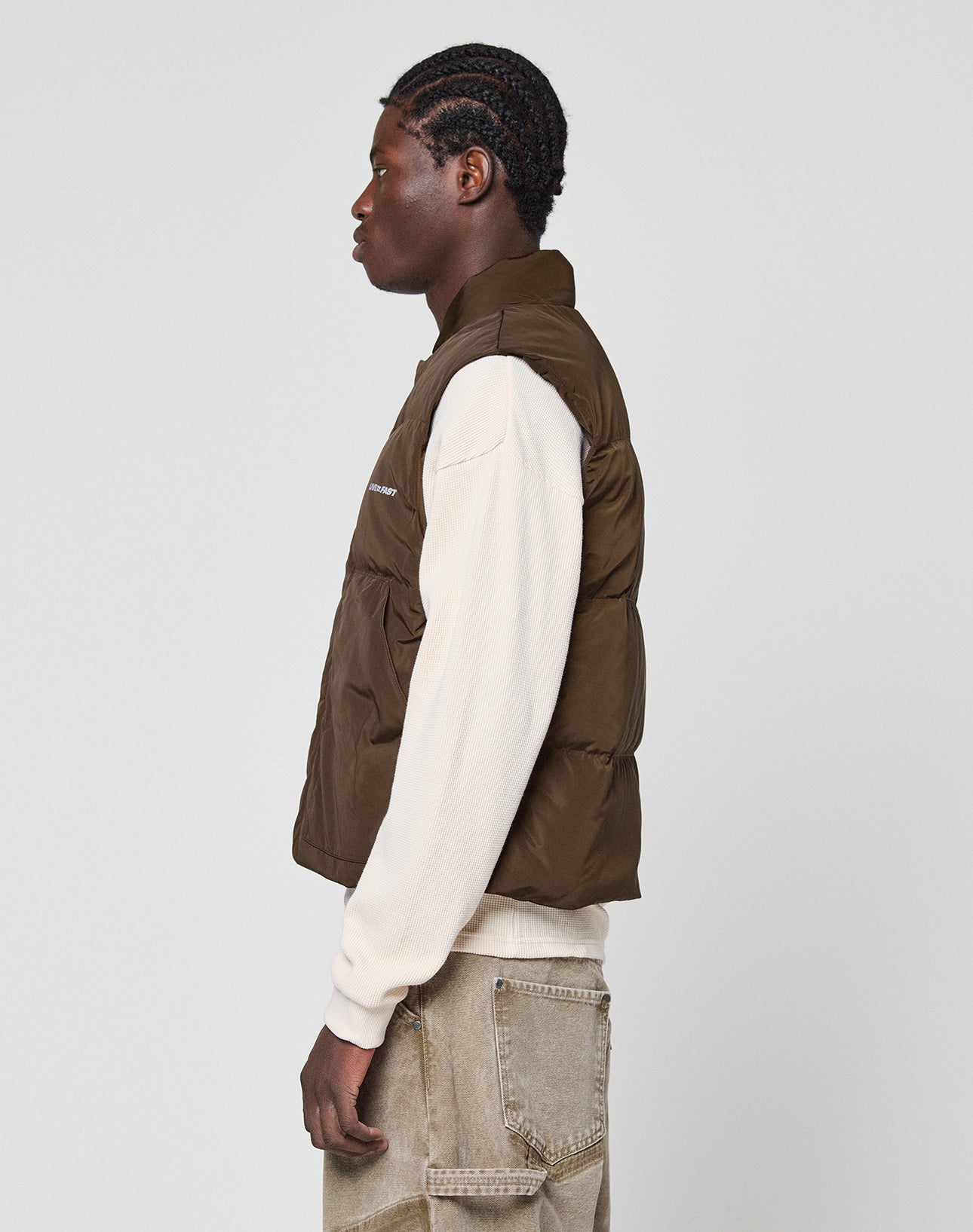 A person with braided hair stands in profile, wearing a cream long-sleeve shirt, light brown jeans, and the LIVE FAST DIE YOUNG Live Fast Cropped Vest in brown polyester, against a plain light gray background.