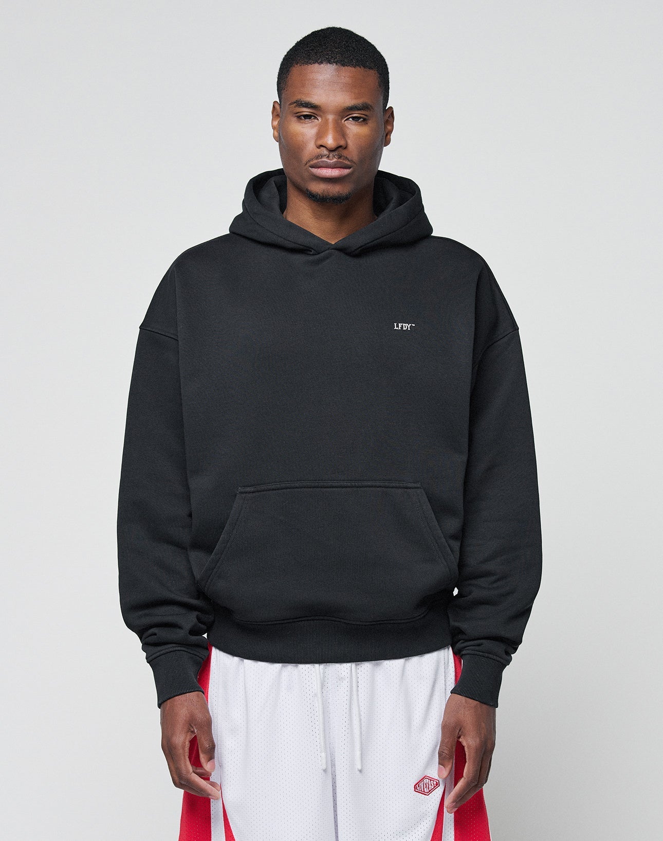 A person stands against a plain background, wearing the LIVE FAST DIE YOUNG Basic Trademark Hooded sweatshirt in black, featuring an oversize fit, small chest logo, and made from 65% Baumwolle, paired with white athletic shorts with red stripes.