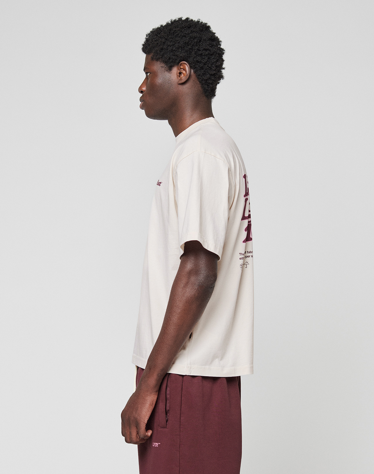 A person stands in profile facing left, wearing the LIVE FAST DIE YOUNG Basic 210 Tee in oversize cream with maroon front and back graphics and maroon shorts, set against a plain light gray background.