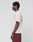 A person stands in profile facing left, wearing the LIVE FAST DIE YOUNG Basic 210 Tee in oversize cream with maroon front and back graphics and maroon shorts, set against a plain light gray background.