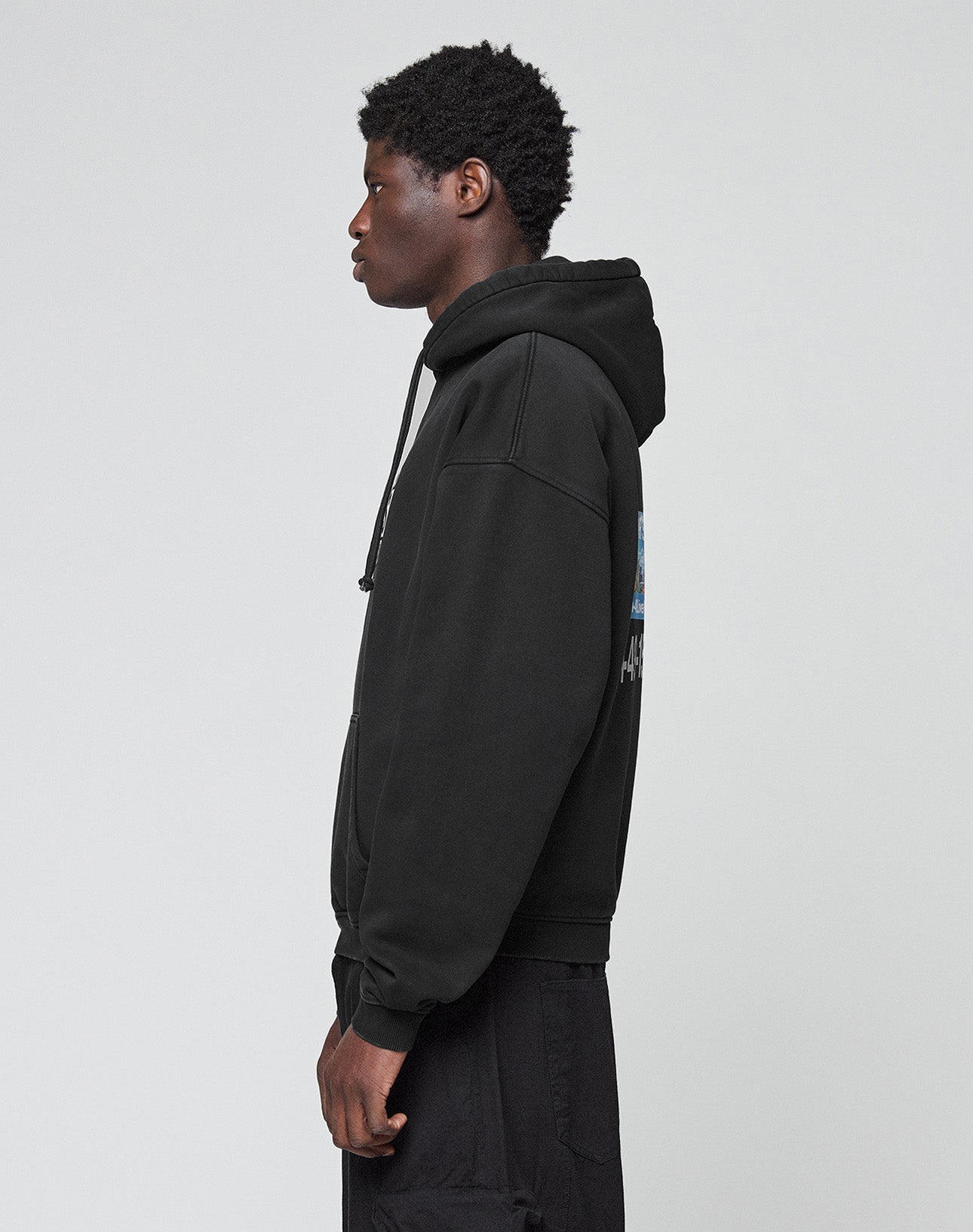 A person with short curly hair stands in profile, wearing the LIVE FAST DIE YOUNG Live Fast Call Us Hooded sweatshirt and black pants made from soft cotton, set against a plain light gray background.