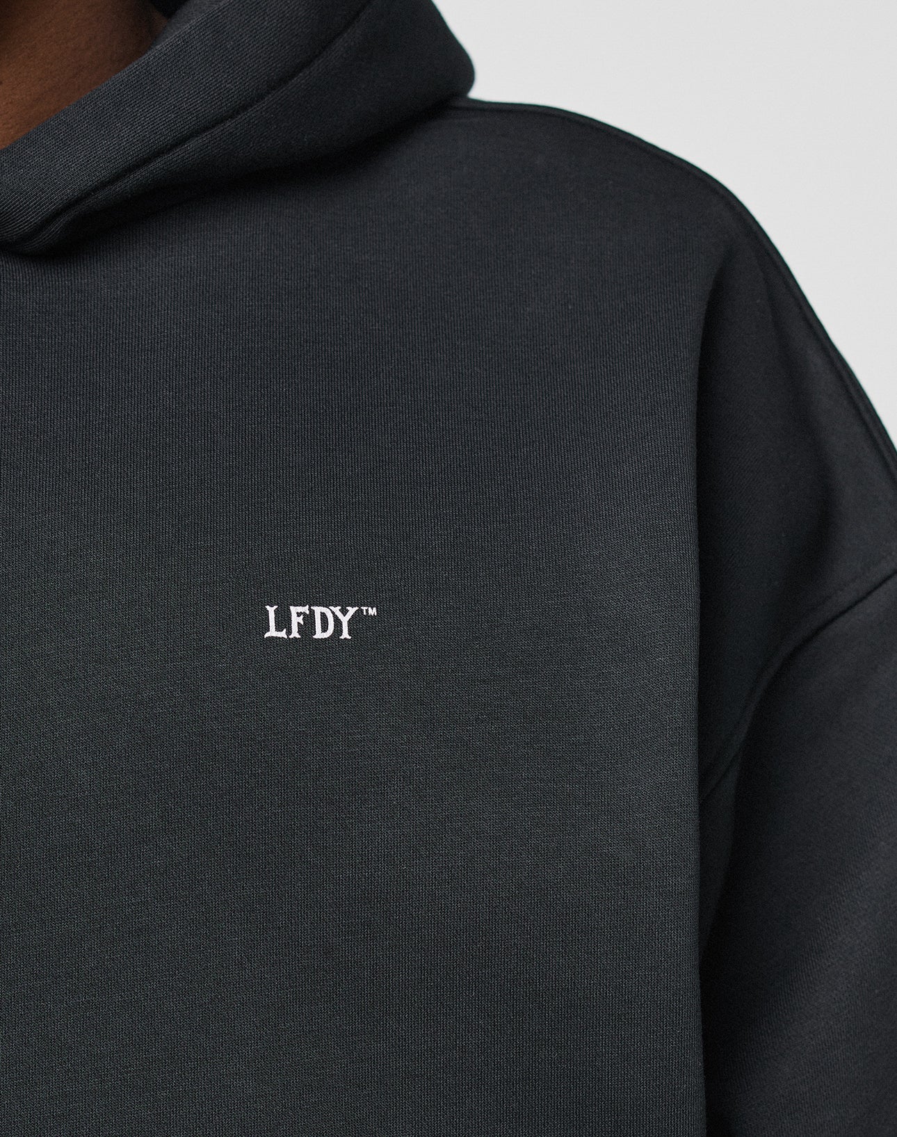 Close-up of a person wearing the Basic Trademark Hooded by LIVE FAST DIE YOUNG, featuring white LFDY™ embroidery on the chest. Made from 65% Baumwolle in an oversize fit for comfort and style. Hood and shoulder seams are visible.