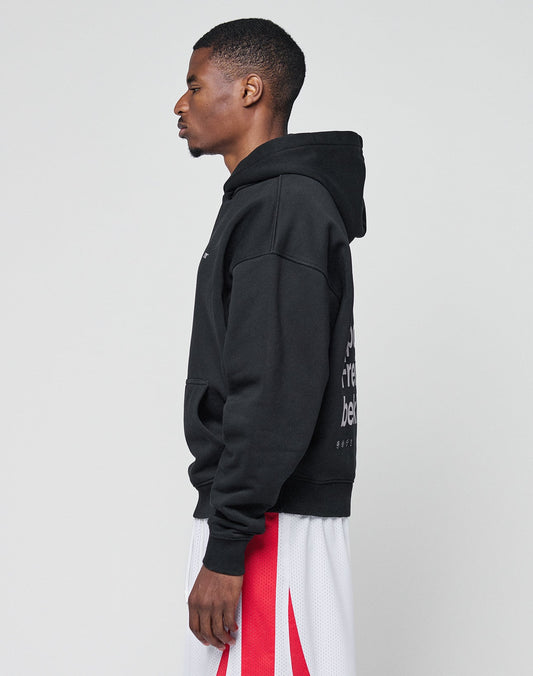 A man stands in profile against a plain background, wearing the LIVE FAST DIE YOUNG Basic Trademark Hooded in black with bold front and back prints, along with white athletic shorts featuring red accents. The hoodie is made from 65% Baumwolle.