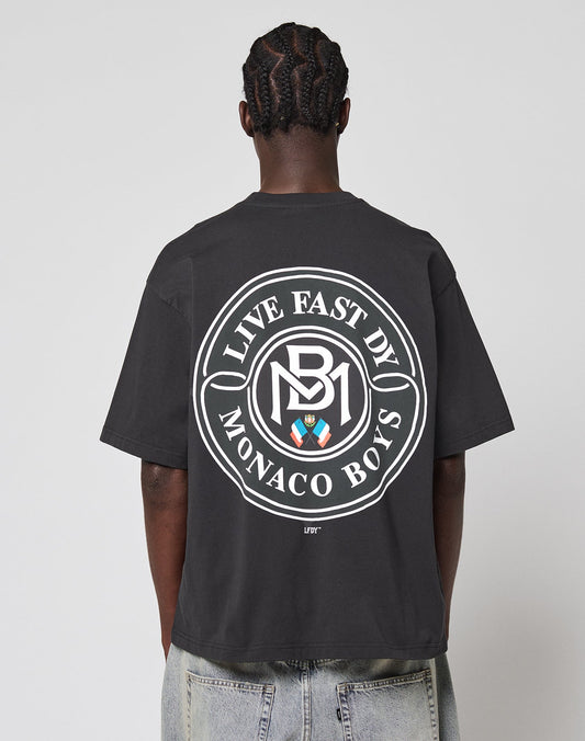 A person with braided hair, facing away, wears the LFDY Monaco Boys Tee—an oversized black T-shirt with a vintage effect and a large circular white LIVE FAST D1 MONACO BOYS logo plus colored emblem on the back.