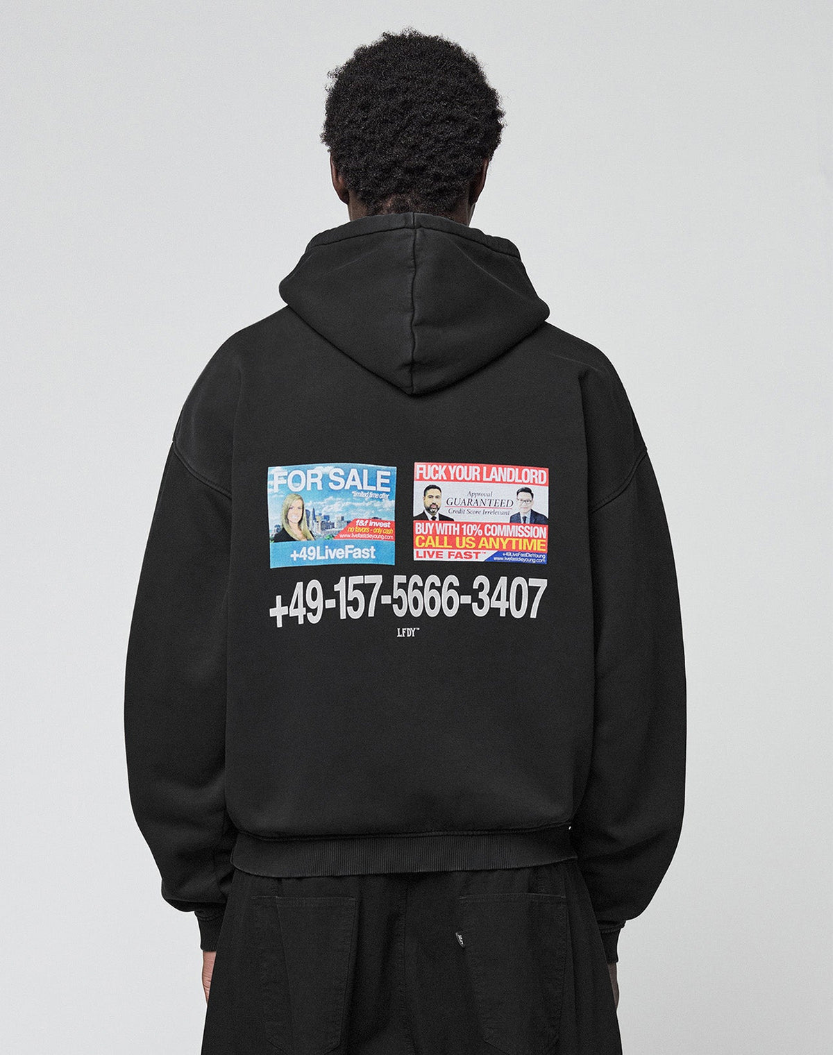 A person is seen from behind wearing the LIVE FAST DIE YOUNG Live Fast Call Us Hooded, an oversize black hoodie made of cotton with bold white text, a phone number, and colorful "For Sale" graphics on front and back, against a plain light background. | model