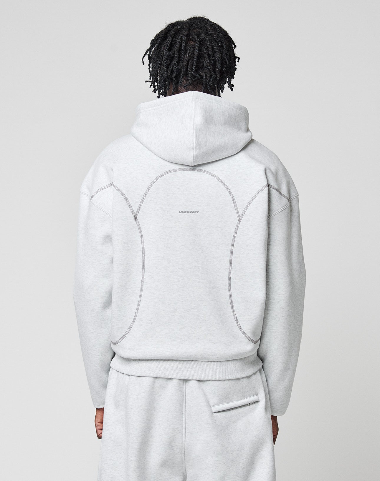 A person with short, twisted hair faces away wearing a LIVE FAST DIE YOUNG LFDY Active Fleece Hooded Zip and matching sweatpants in light gray, featuring curved seams and Logo Print text on the back against a plain background.