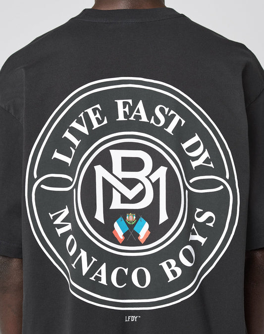 Someone wearing the LFDY Monaco Boys Tee: an oversized black shirt with a large circular white graphic on the back reading LIVE FAST DY MONACO BOYS, featuring a crest, flags, and MB initials. Vintage effect, 100% cotton.