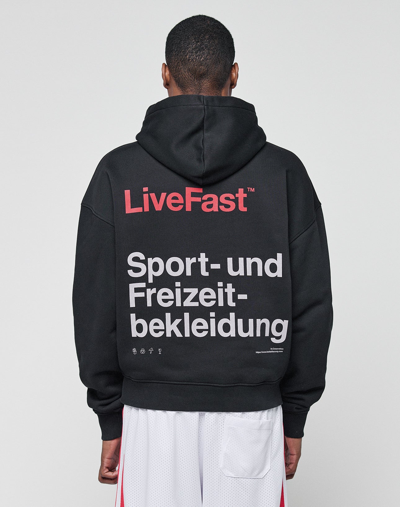 A person with short hair, seen from behind, wears the LIVE FAST DIE YOUNG Basic Trademark Hooded in black with red LiveFast and white Sport- und Freizeitbekleidung text front and back, paired with white athletic shorts on a plain gray background.