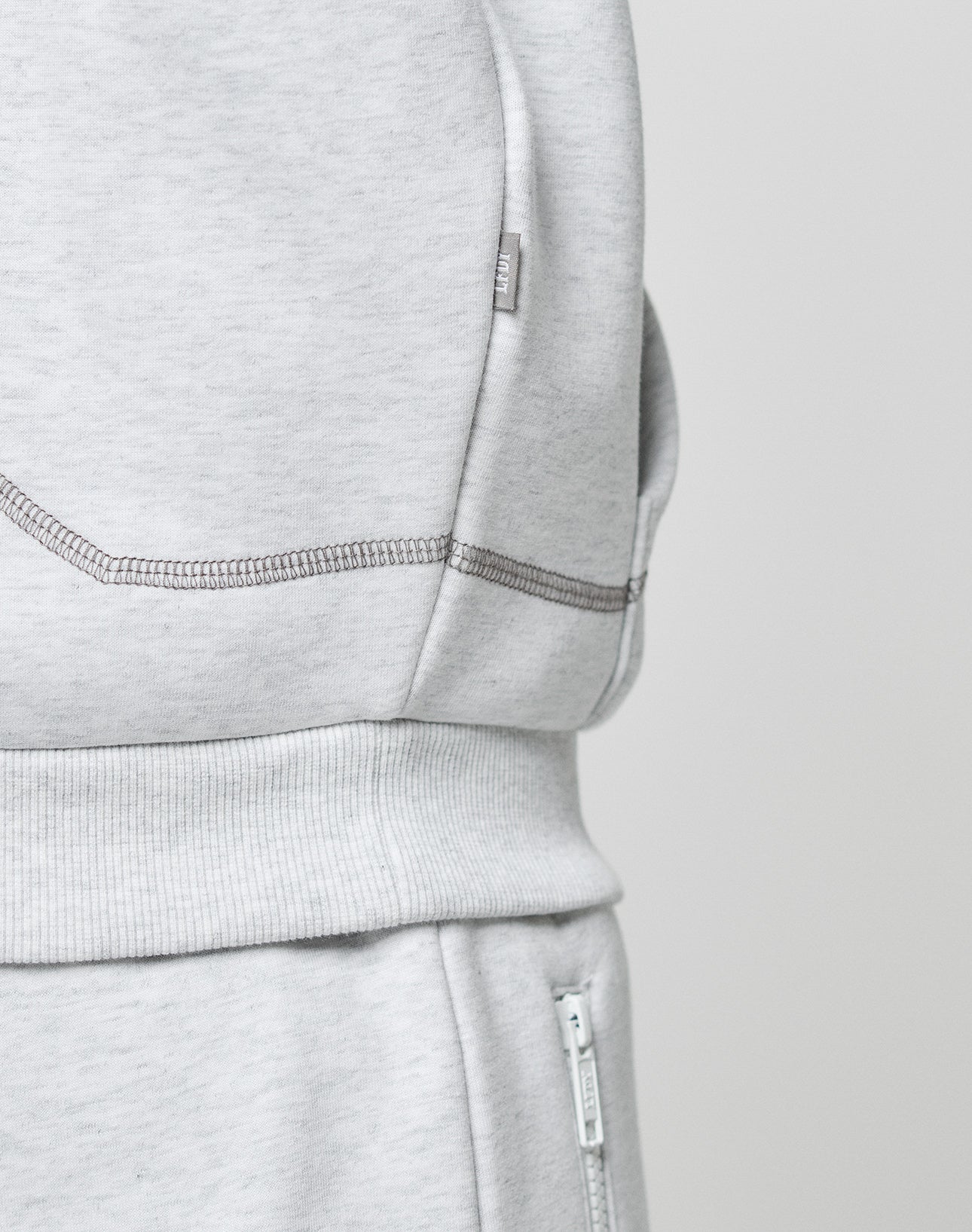 Close-up of the LFDY Active Fleece Hooded Zip set by LIVE FAST DIE YOUNG, light gray with detailed side pocket stitching, logo fabric tag, and a visible zipper on the oversized pants’ waistband.