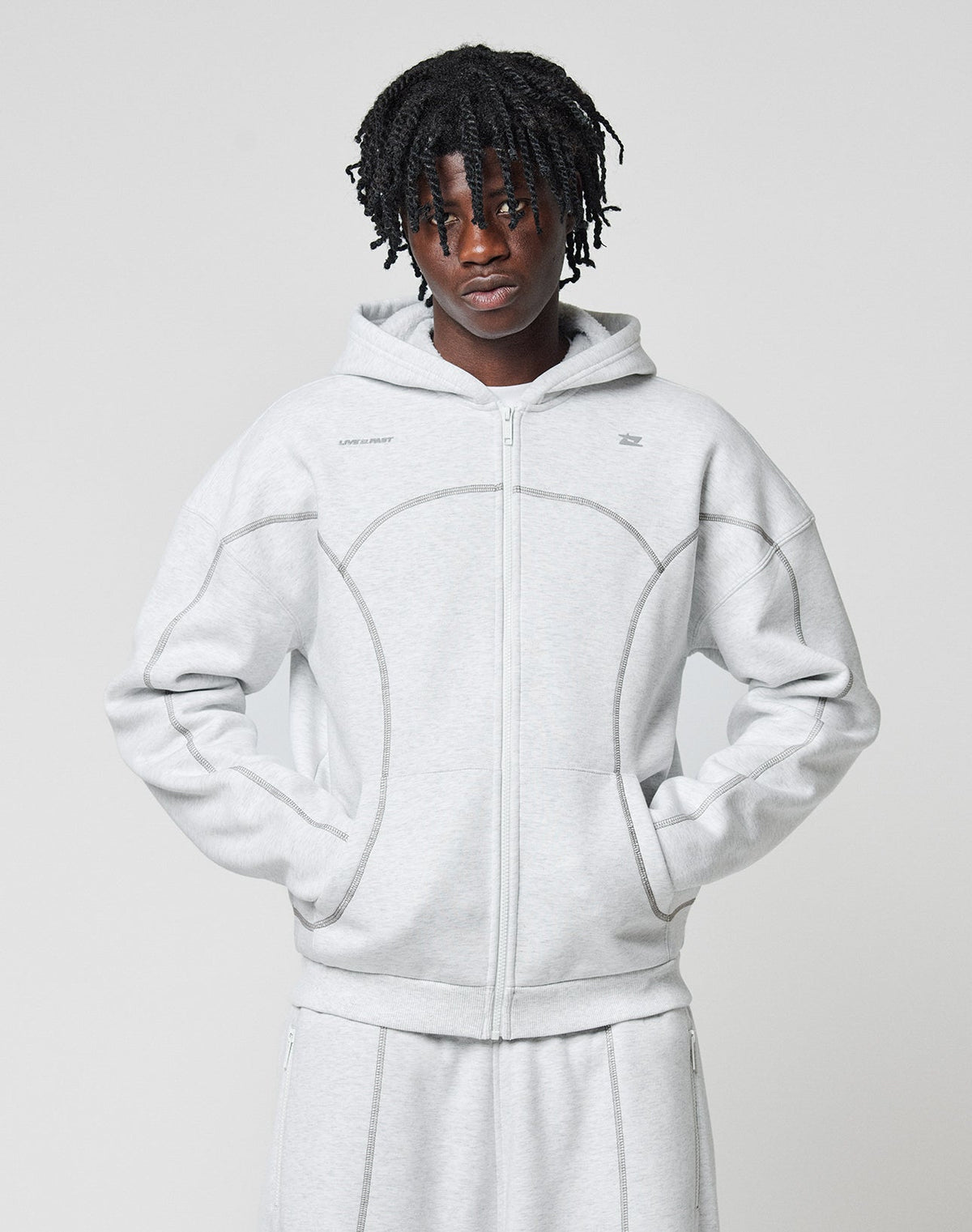 A person with short dreadlocks wears the LIVE FAST DIE YOUNG LFDY Active Fleece Hooded Zip in light gray and matching pants, posing against a plain background with hands in the sweatshirt’s pockets.