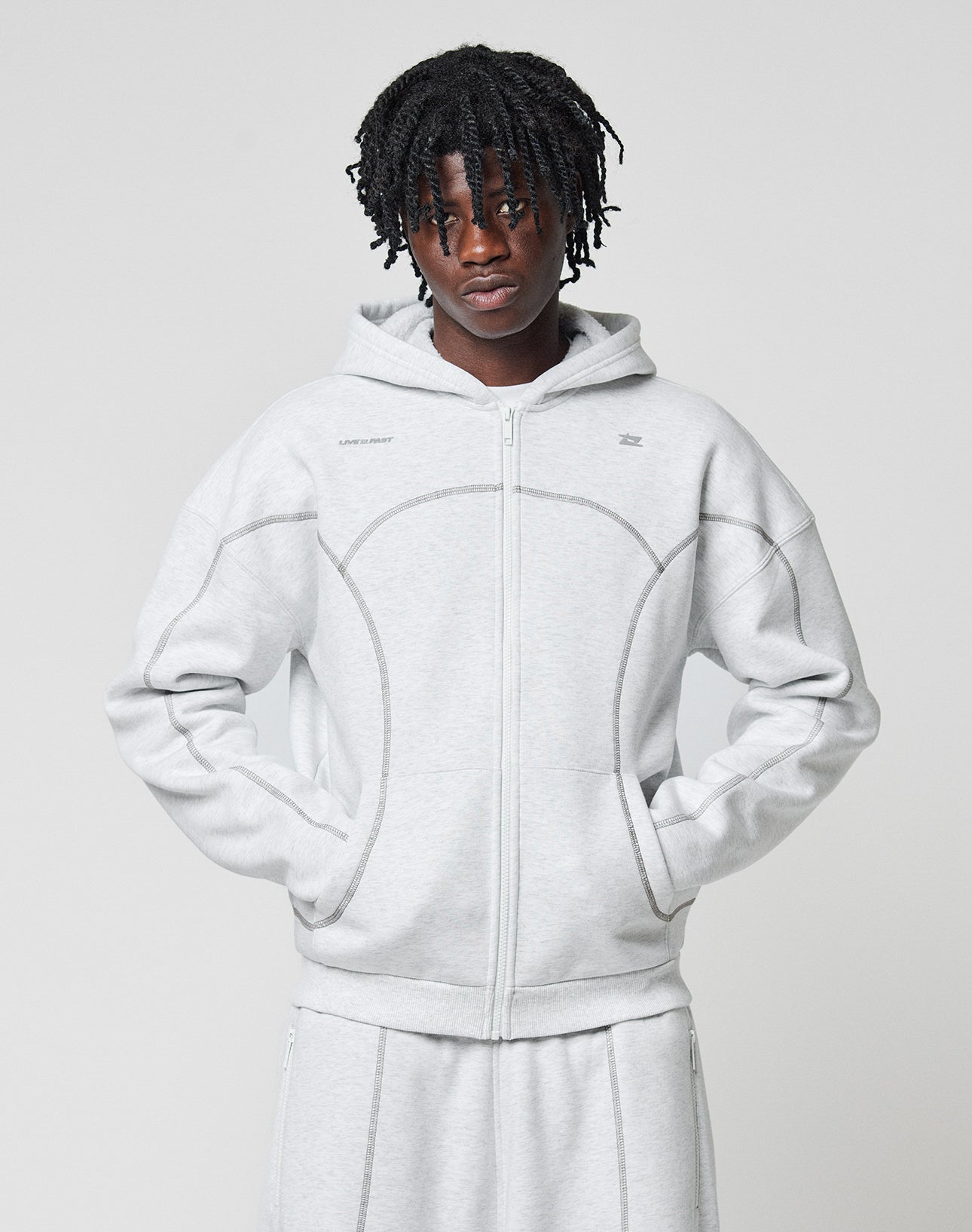 A person with short dreadlocks wears the LIVE FAST DIE YOUNG LFDY Active Fleece Hooded Zip in light gray and matching pants, posing against a plain background with hands in the sweatshirt’s pockets.