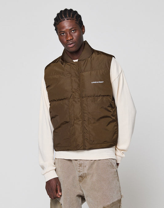 A man wears the LIVE FAST DIE YOUNG Live Fast Cropped Vest, a regular fit brown polyester puffer, over a cream sweatshirt and light brown jeans, standing against a plain light gray background and looking at the camera neutrally.