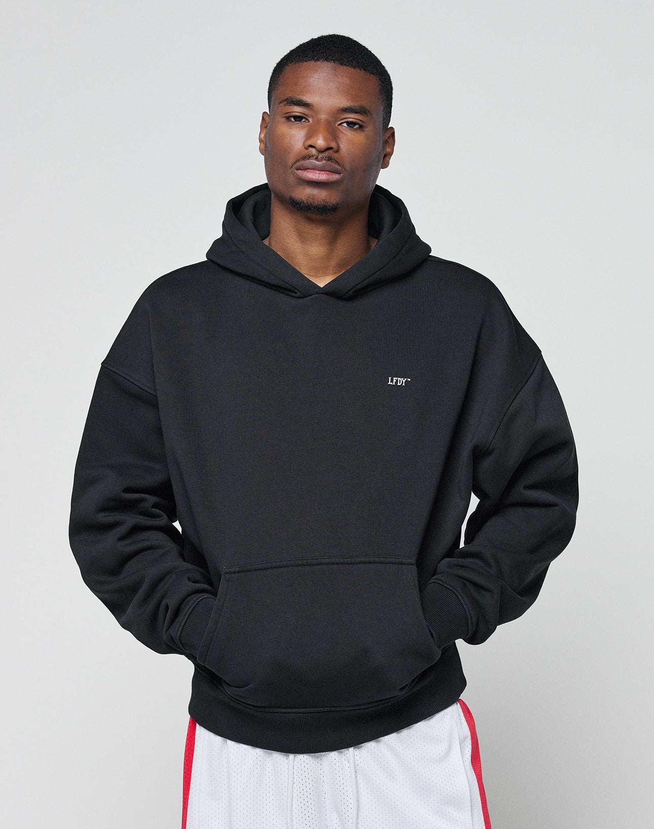 A man stands against a plain background wearing the LIVE FAST DIE YOUNG Basic Trademark Hooded in black (65% Baumwolle) with LFYT printed on the chest and white athletic shorts with red stripes, hands in his hoodie pocket and a neutral expression.