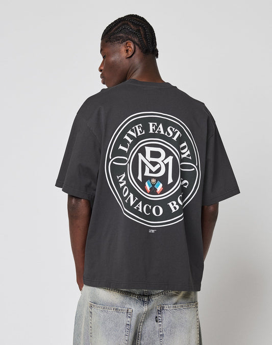 A person with braided hair and light denim jeans faces away, wearing the LFDY Monaco Boys Tee by LIVE FAST DIE YOUNG in oversized fit, vintage optic, featuring a large circular back graphic with LIVE FAST, DIE YOUNG MONACO BOYS text.