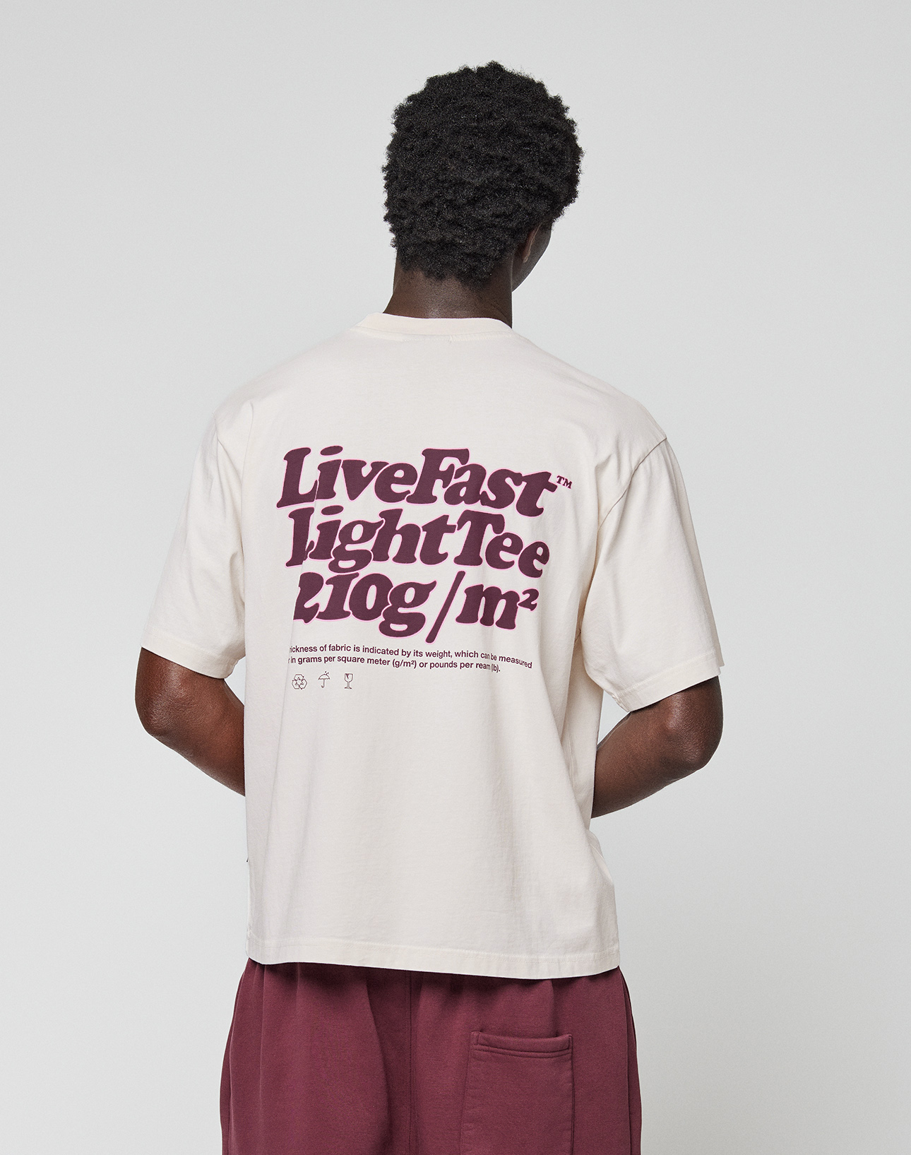 A person faces away, wearing an oversized cream Basic 210 Tee by LIVE FAST DIE YOUNG, made from 100% Baumwolle with a bold maroon back print and small text. The look is finished with maroon shorts.