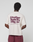 A person faces away, wearing an oversized cream Basic 210 Tee by LIVE FAST DIE YOUNG, made from 100% Baumwolle with a bold maroon back print and small text. The look is finished with maroon shorts.