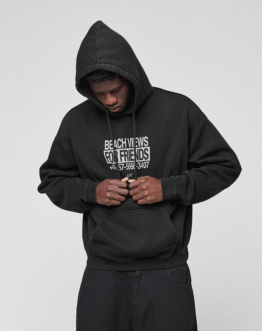 A person in an Oversize Fit black Live Fast Call Us Hooded by LIVE FAST DIE YOUNG, hood up and hands in the kangaroo pocket, looks down. The cotton hoodie features white Beach Views For Friends text and a phone number underneath.
