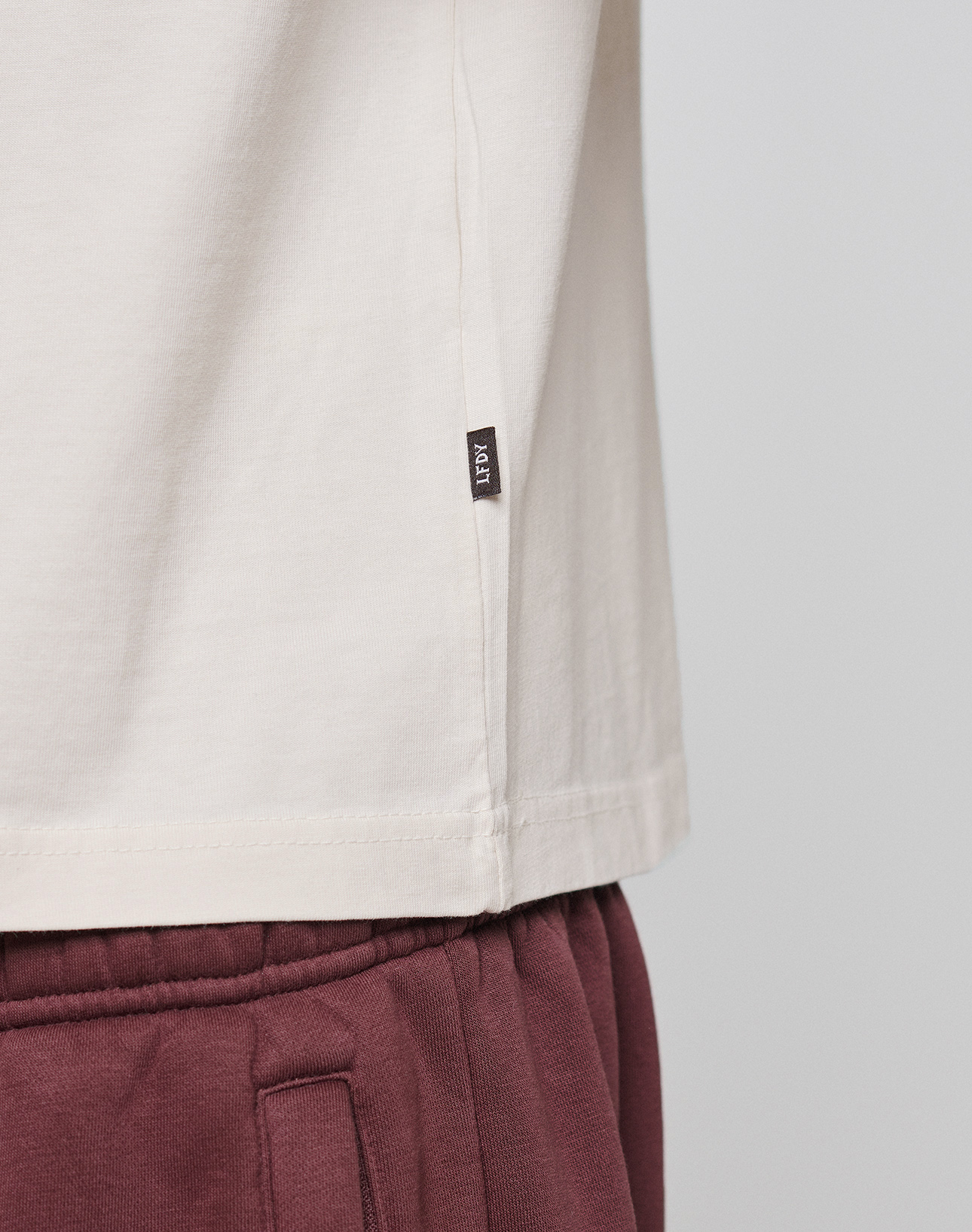 A close-up of a person wearing the LIVE FAST DIE YOUNG Basic 210 Tee in cream (Oversize Fit) with a small black ICON tag on the side seam, paired with maroon pants. Visible are only the shirt’s lower part and top of the pants.