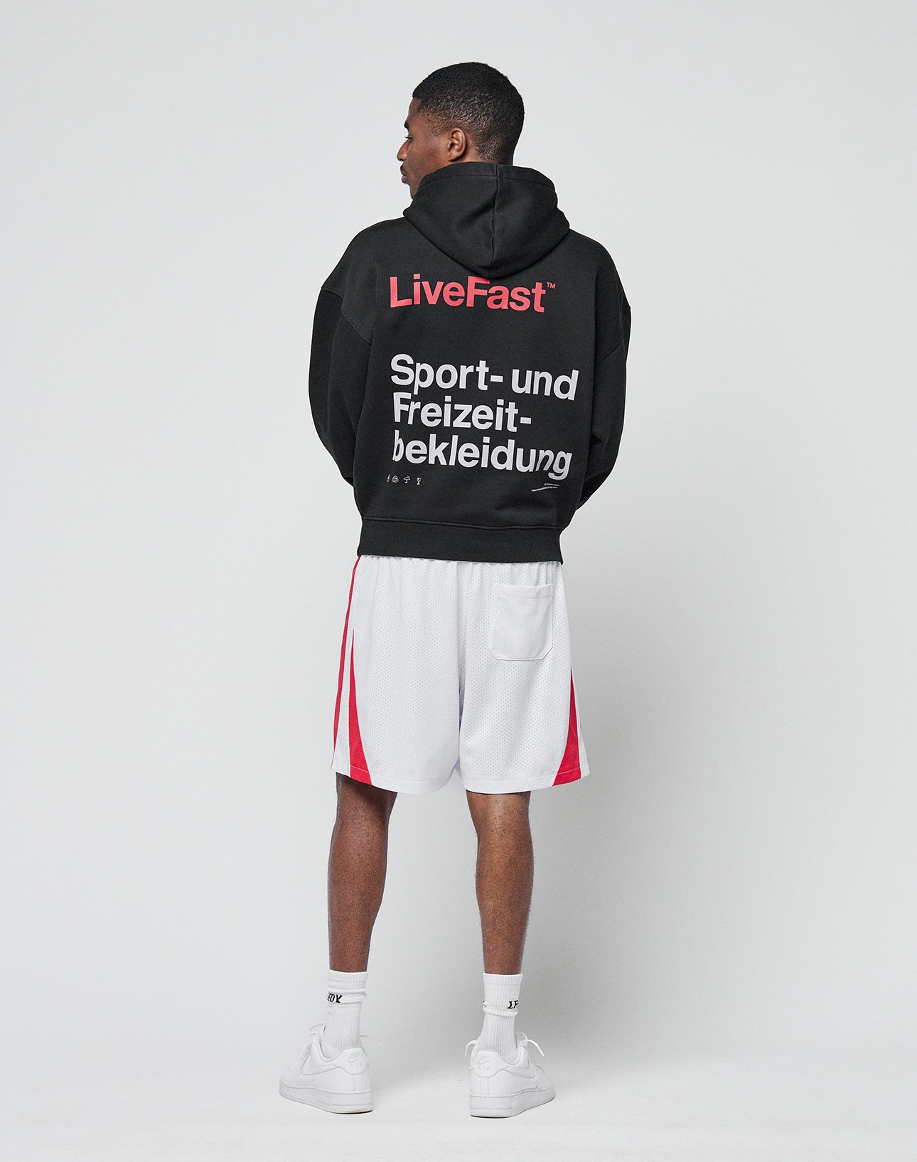 A man faces away, wearing the LIVE FAST DIE YOUNG Basic Trademark Hooded in black with white and red Front- und Backprint German text, paired with white athletic shorts with red stripes, white socks, and sneakers against a plain light background.