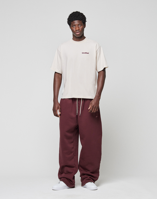 A person stands against a plain background wearing the LIVE FAST DIE YOUNG Basic 210 Tee in oversize beige with front and back Livefast print, paired with loose maroon drawstring sweatpants and white sneakers.