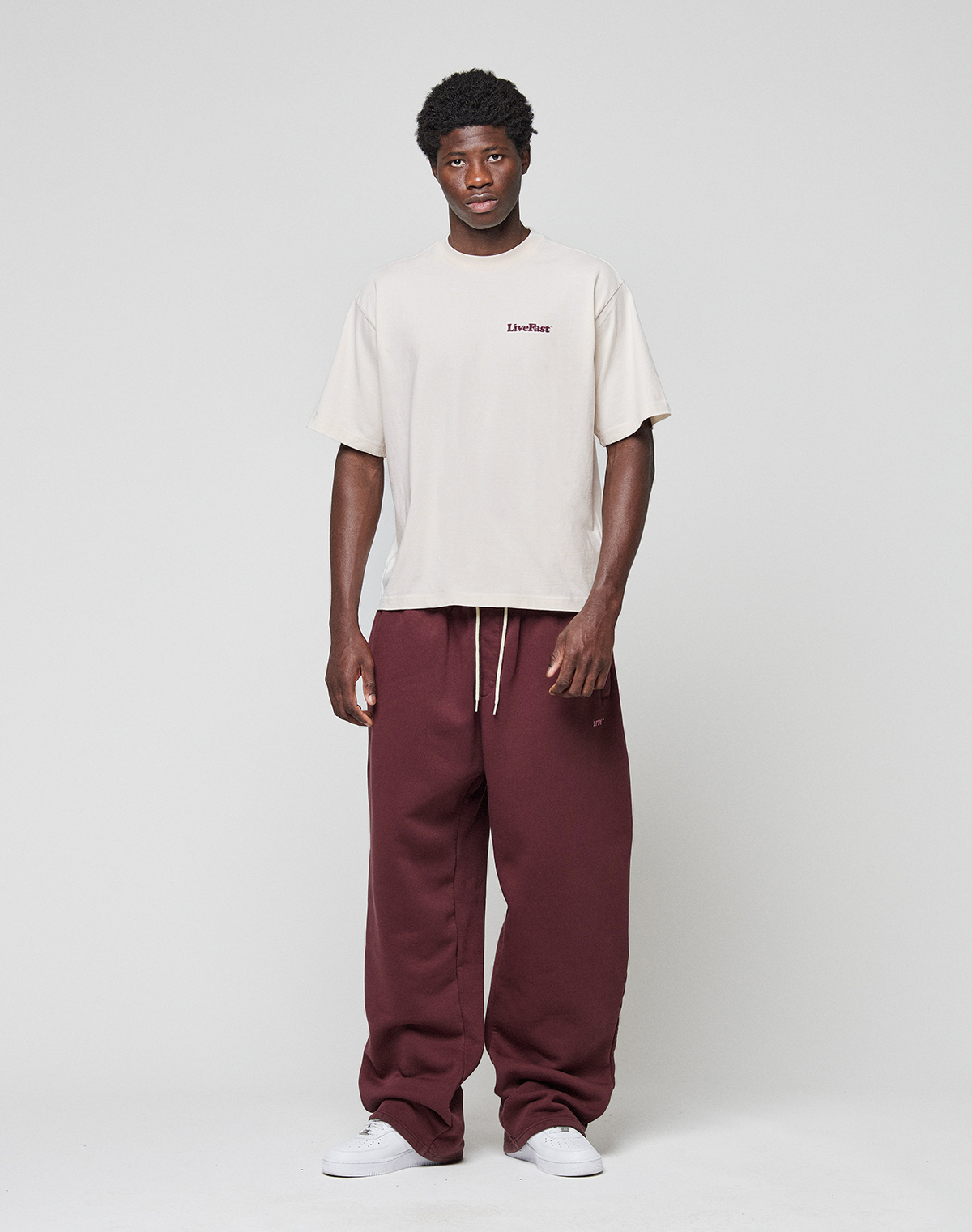 A person stands against a plain background wearing the LIVE FAST DIE YOUNG Basic 210 Tee in oversize beige with front and back Livefast print, paired with loose maroon drawstring sweatpants and white sneakers.