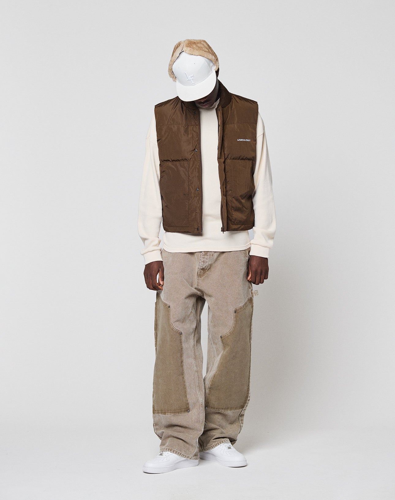 Wearing a white cap, cream sweatshirt, Live Fast Cropped Vest by LIVE FAST DIE YOUNG, loose tan patchwork jeans, and white sneakers, a person stands against a plain light background, looking down with hands by their sides.