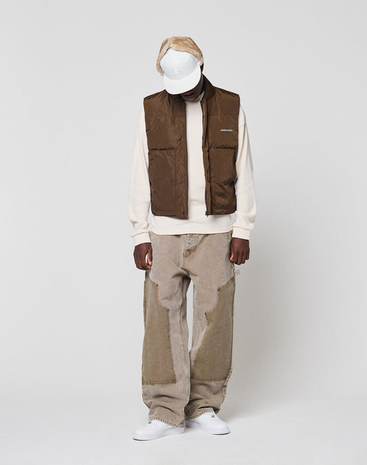 Wearing a white cap, cream sweatshirt, Live Fast Cropped Vest by LIVE FAST DIE YOUNG, loose tan patchwork jeans, and white sneakers, a person stands against a plain light background, looking down with hands by their sides.