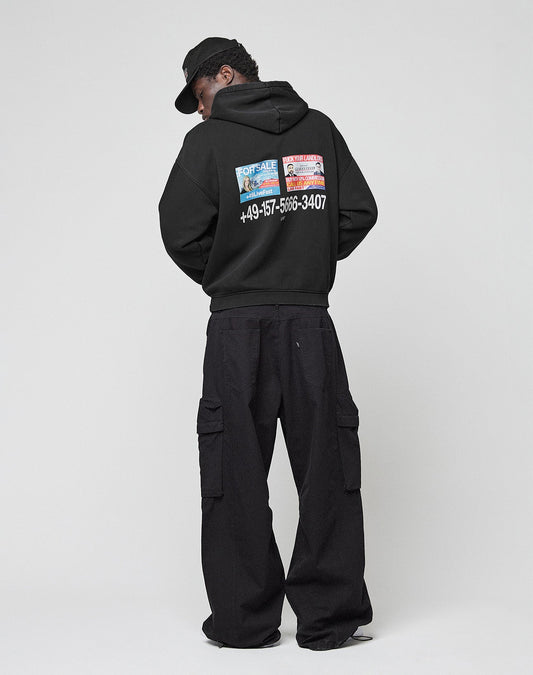 A person wearing the LIVE FAST DIE YOUNG Live Fast Call Us Hooded, an oversize black cotton hoodie with colorful prints, black cargo pants, and a cap stands facing away against a plain light background.