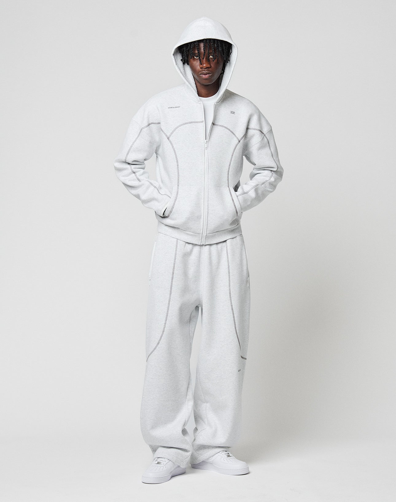 A person faces forward against a plain background, wearing the LIVE FAST DIE YOUNG LFDY Active Fleece Hooded Zip in light gray with matching sweatpants and white sneakers. The hood is up and hands are in pockets.