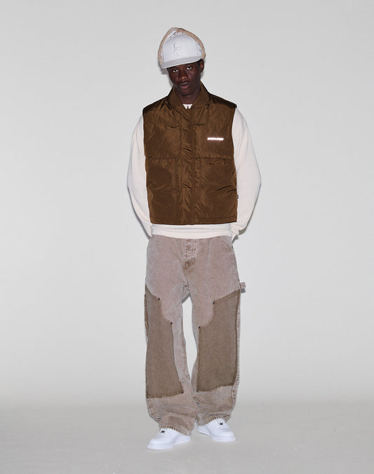 A person stands against a plain light background wearing the LIVE FAST DIE YOUNG Live Fast Cropped Vest in brown over a cream long-sleeve shirt, beige patchwork pants, a beige hat, and white sneakers.
